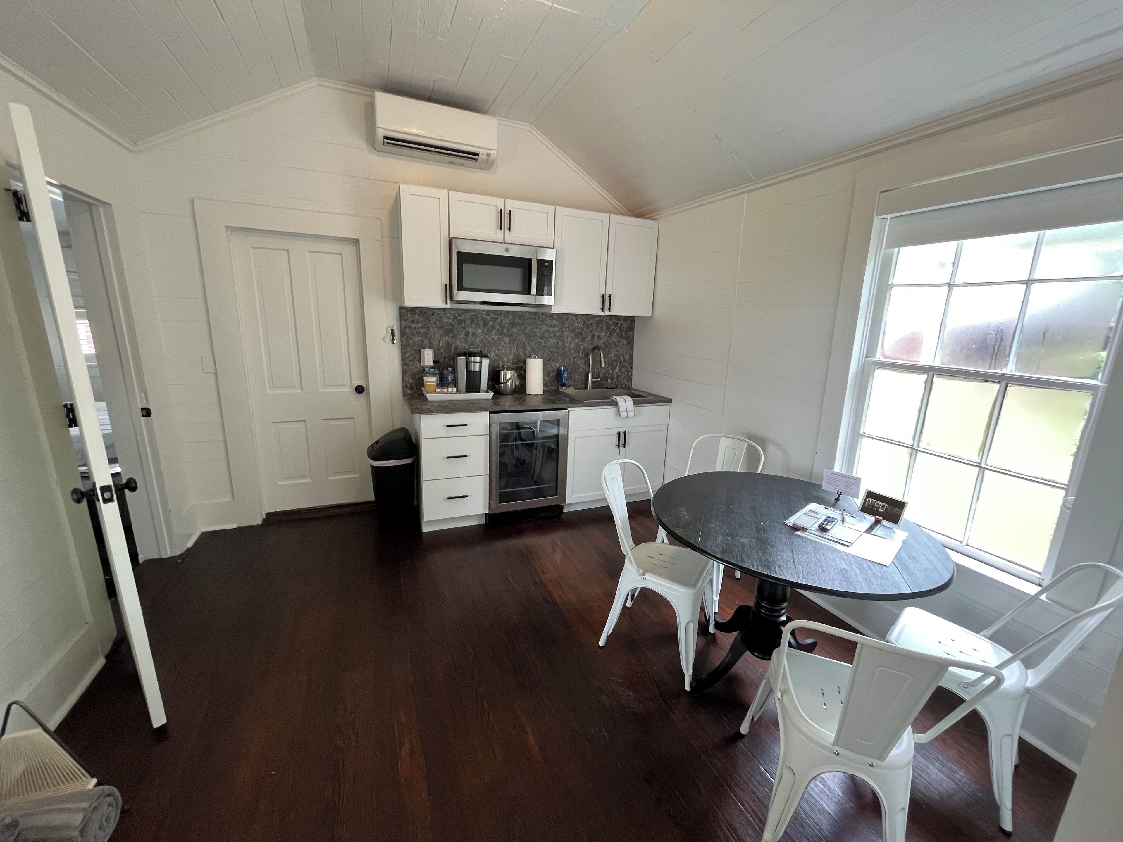 A compact kitchen with white cabinets, a microwave, and a sink. There's a dining table with chairs and a window with a view.