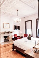 Een slaapkamer met een groot bed, een open haard, een kroonluchter, een rode fauteuil en een televisie aan de muur.