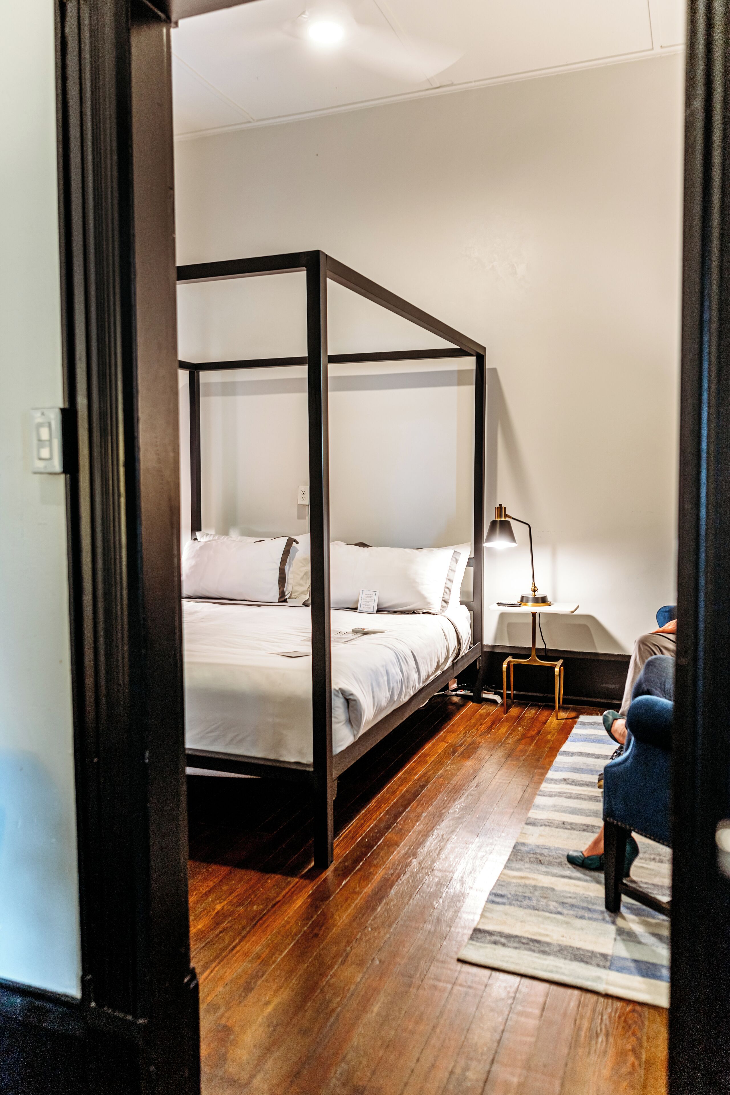 A four-poster bed in a room with wooden flooring, a bedside table with a lamp, and a striped rug.