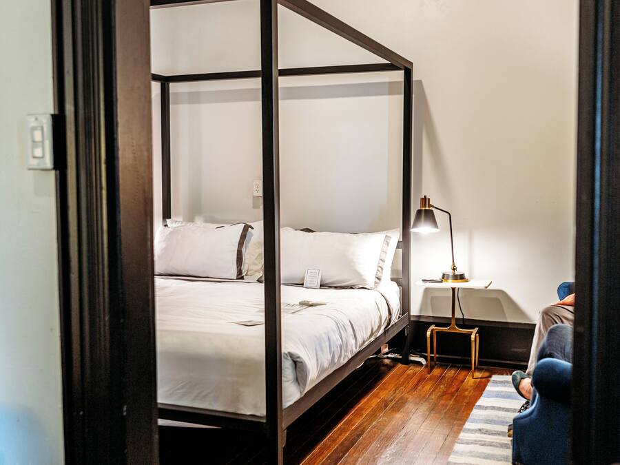 A four-poster bed in a room with wooden flooring, a bedside table with a lamp, and a striped rug.