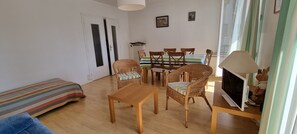 A living room with a sofa, a wooden table, wicker chairs, a striped rug, a dining table with chairs, a TV, and a lamp. - Beautiful apartment in the center of Concarneau with private parking (Concarneau)