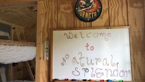 Eine Tafel mit der Aufschrift 'Welcome to Natural Splendor' in bunten Buchstaben, eine Uhr mit Basketball und Hornets-Logo sowie ein hölzernes Etagenbett.
