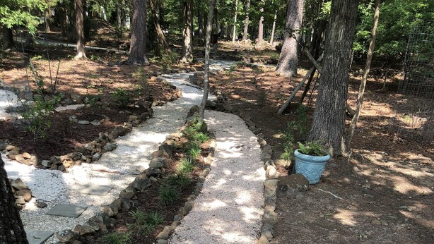 Ein Kiesweg, der von Steinen und kleinen Pflanzen gesäumt wird, führt durch ein bewaldetes Gebiet mit Bäumen und einem Zaun im Hintergrund.