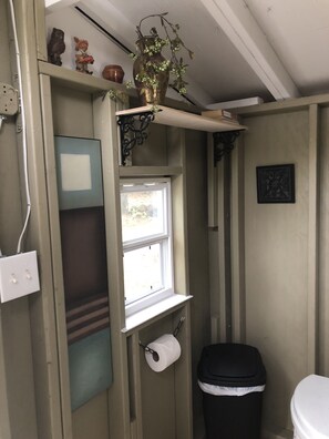 Un baño pequeño con ventana, inodoro, cubo de basura y estante con adornos.