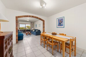 A dining area with a wooden table and chairs, a sofa, and a view through an archway to a living room with a couch and a dining table.