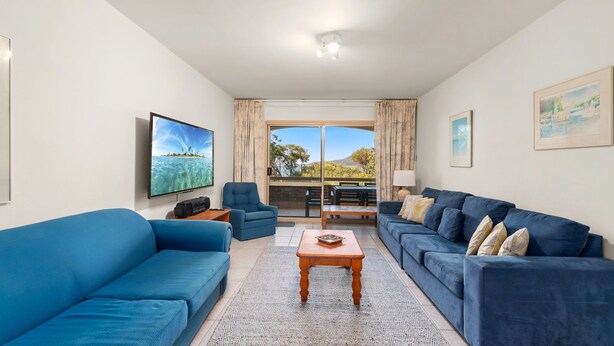 A living room with a large TV, two blue sofas, a wooden coffee table, and a view of the outdoors through a sliding glass door.