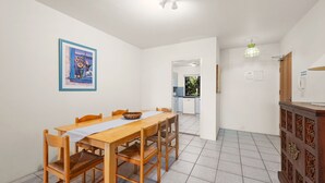 A dining area with a wooden table and chairs, a framed picture on the wall, and a kitchen in the background.