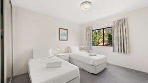 A room with two single beds, a small bedside table with a lamp, a framed picture on the wall, and a window with curtains.