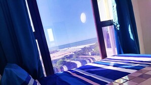 A bed with a striped blue and white bedspread, a pillow, and a view of a beach and buildings through a window with blue curtains. - Claridges 104 (Margate)