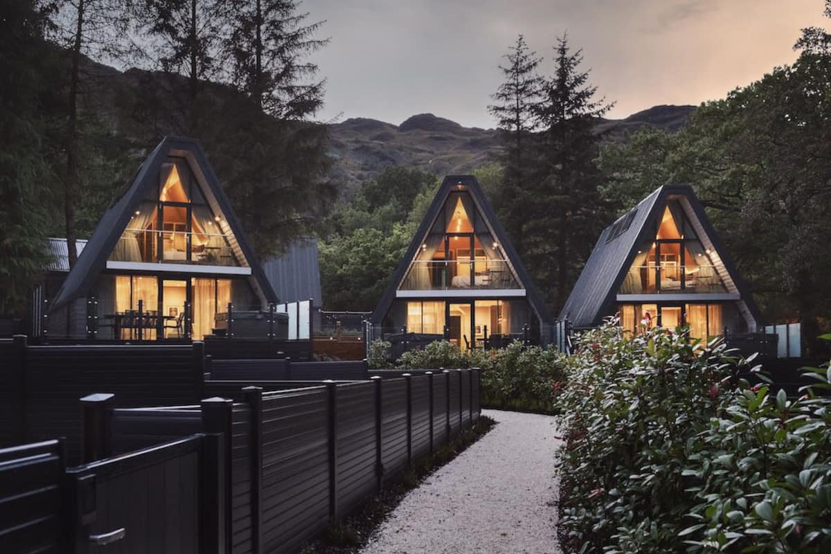 A modern, triangular-roofed cabin with a visible interior, surrounded by dense forest and a gravel path.