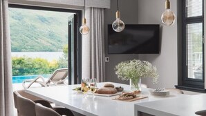 A modern kitchen with a large island, pendant lights, a flat-screen TV, and a view of a lake. - Ardlui Retreat (Arrochar)
