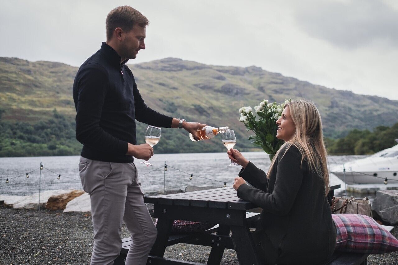 A couple toasting with wine glasses at a wooden table by a lake with mountains in the background.
