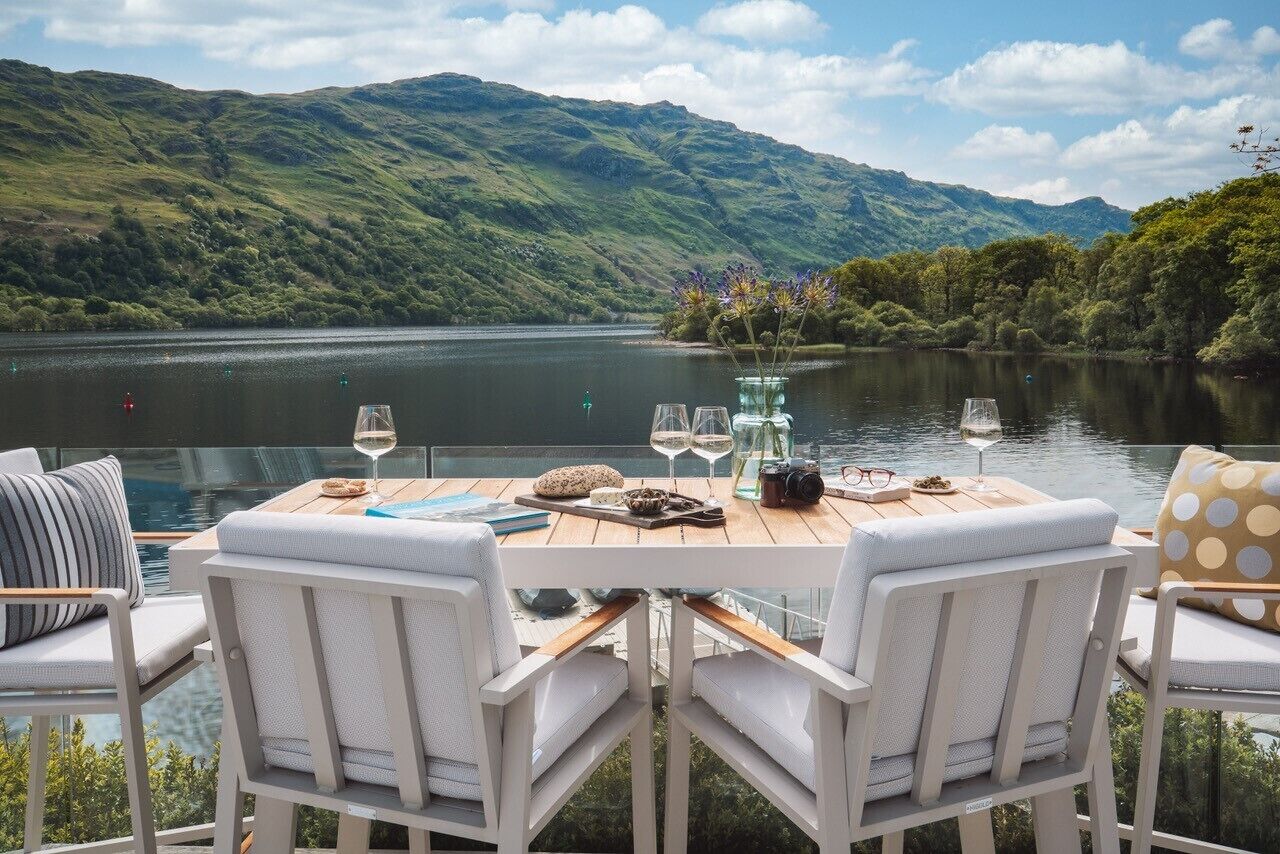 Une table à manger avec des chaises, un vase rempli de fleurs et des verres à vin sur une terrasse donnant sur un lac, avec des collines en arrière-plan.