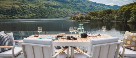 Une table à manger avec des chaises, un vase rempli de fleurs et des verres à vin sur une terrasse donnant sur un lac, avec des collines en arrière-plan.