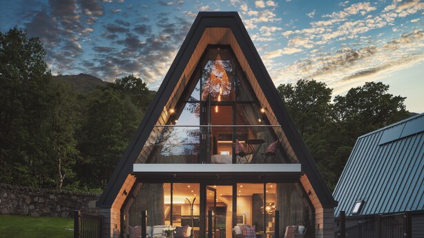 A modern, triangular-shaped building with large glass windows, a cozy interior with seating, and a stone wall in the background. - Ardlui Retreat (Arrochar)
