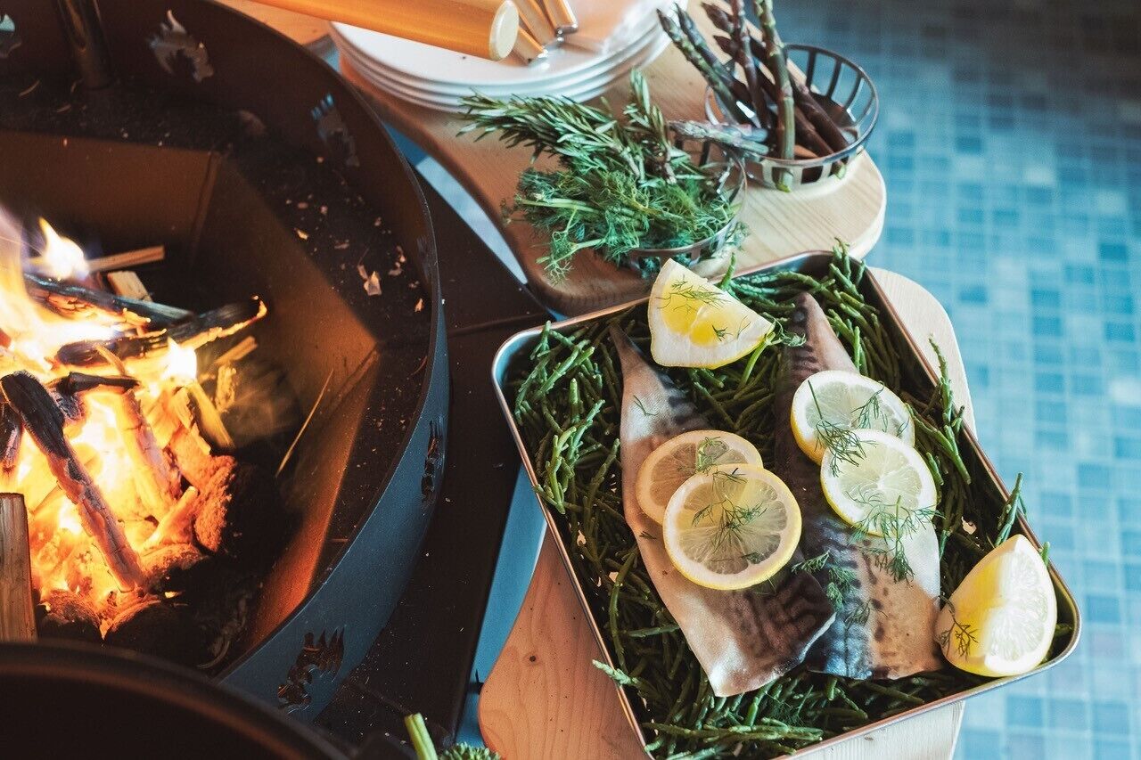 A grill with wood burning, a tray of fish garnished with lemon slices and herbs, and a tiled floor in the background.
