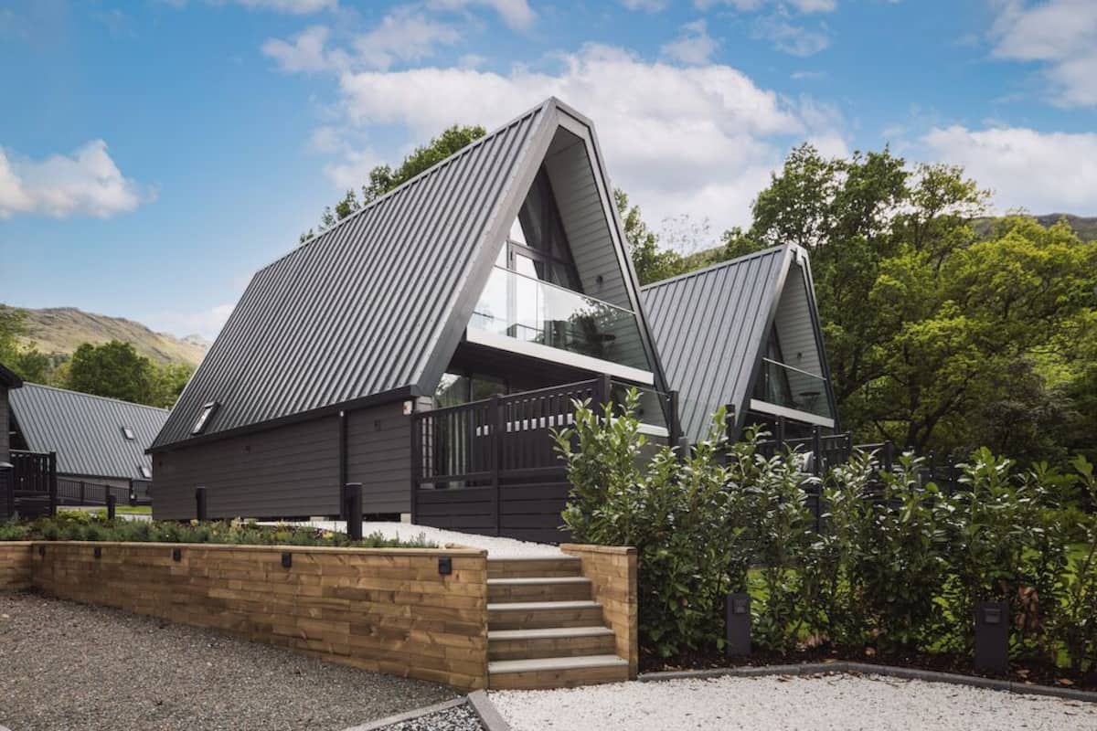 A modern house with a sloped roof, a wooden staircase leading to a glass door, and a view of the surrounding greenery.