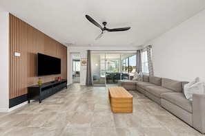 Uma sala de estar moderna com uma televisão de tela plana, um ventilador de teto, uma mesa de centro de madeira, um sofá bege e uma porta de vidro que dá para uma varanda.