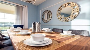 Une salle à manger avec une table en bois dressée pour six personnes, deux miroirs décoratifs accrochés au mur et une vue sur une maison par la fenêtre.