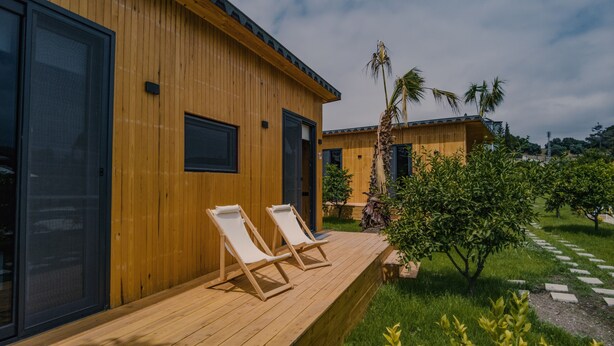 A wooden building with two lounge chairs on a deck, surrounded by greenery and palm trees. - Arsuz Bungalov (Arsuz)