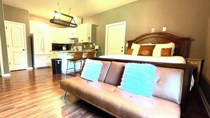 A hotel room with a brown leather sofa, a wooden headboard, a kitchen area, and a ceiling fan. - Lovely Guest Apartment<br> (Pace)