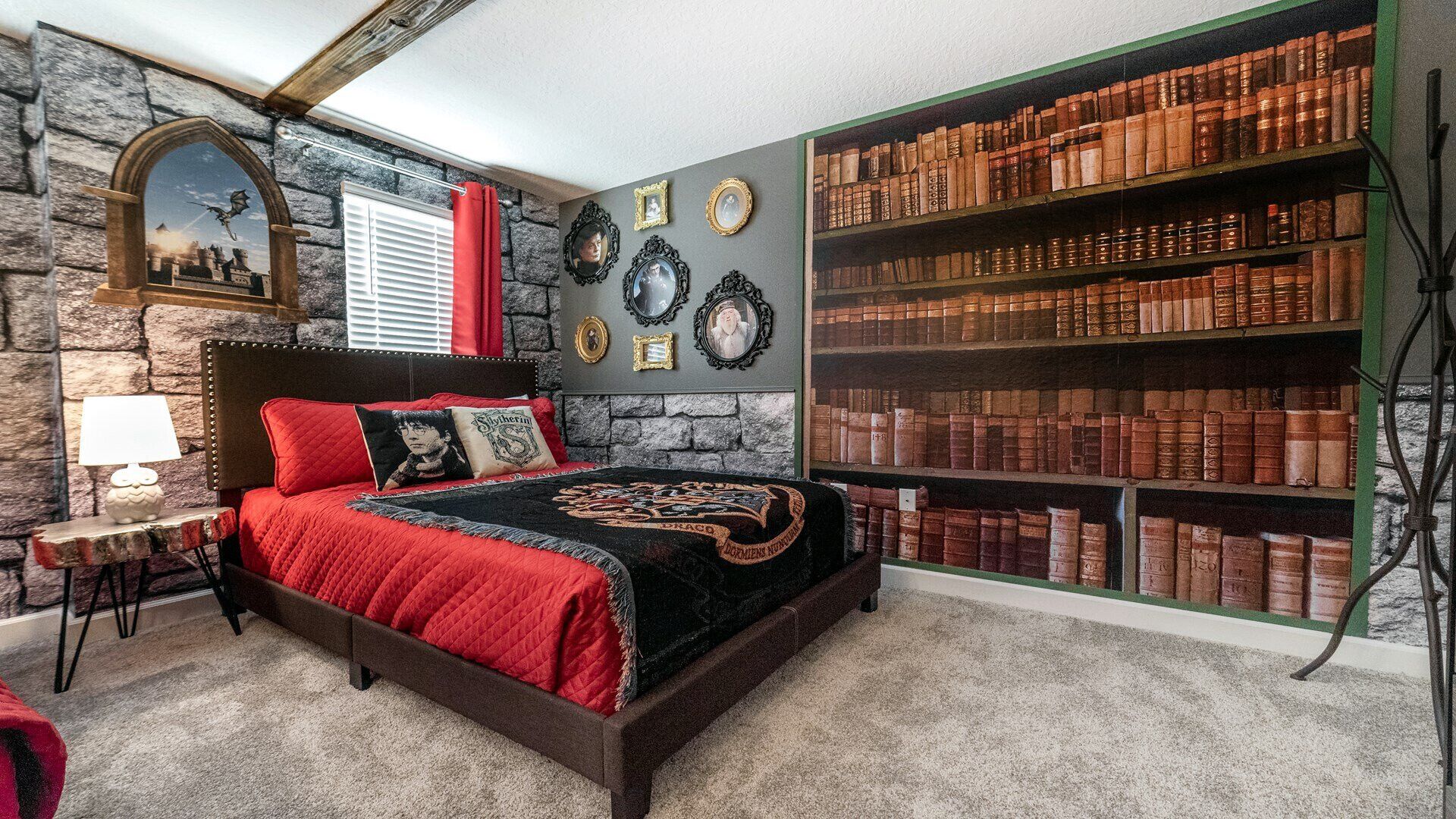 Une chambre avec un mur en pierre, une grande bibliothèque remplie de livres, un lit recouvert d’une courtepointe rouge et un tableau représentant un dragon accroché au mur.