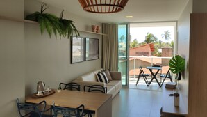 A modern living room with a sofa, dining table, chairs, and a large window with a view of palm trees. - Pé na Areia, Vista Mar e 3 Qts com AC (Barra de São Miguel)
