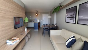 A modern living room with a sofa, a dining area with chairs, a kitchen in the background, and a large plant on the wall. - Pé na Areia, Vista Mar e 3 Qts com AC (Barra de São Miguel)