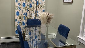 Une salle de conférence avec une table en verre, des chaises bleues, un rideau à motifs floraux, un vase contenant des plantes séchées et un tableau encadré accroché au mur.