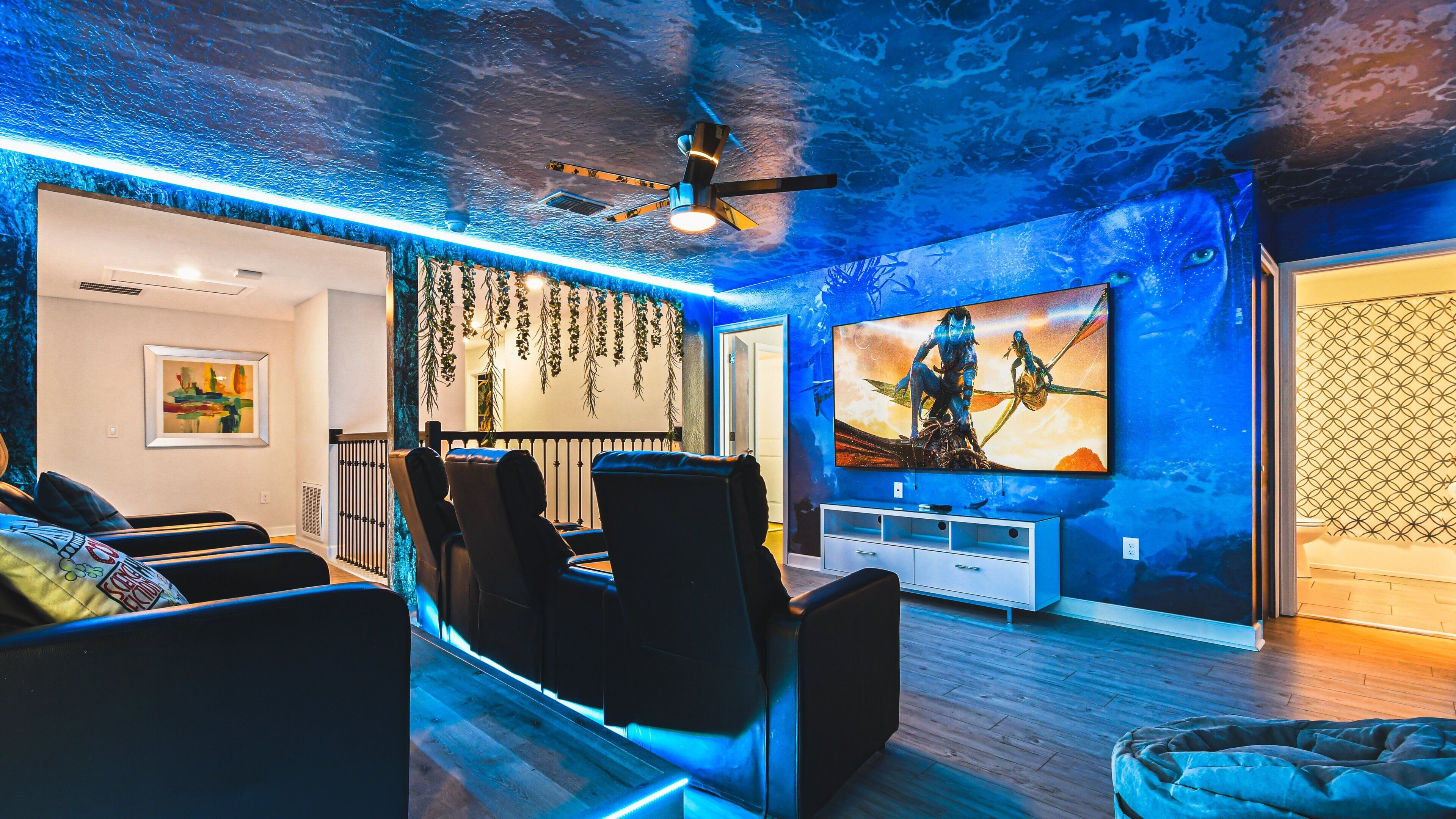 Un salon moderne avec un plafond éclairé de lumière bleue, un grand téléviseur et un ventilateur de plafond.