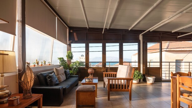 Amplia sala de estar con sofá, sillas de madera, una mesa de centro y plantas en macetas. Grandes ventanas ofrecen vistas al mar y a las montañas.