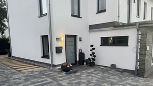 Un bâtiment moderne à la façade épurée et blanche, avec une porte noire et un petit jardin agrémenté d’un pot de fleurs et d’un vase décoratif.