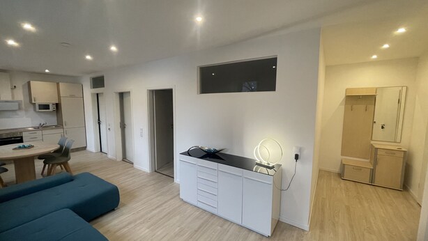 Une pièce moderne et bien éclairée, dotée d’une kitchenette, d’une coiffeuse avec miroir et d’un coin salon avec des canapés bleus.