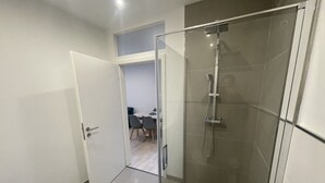 Une salle de bain moderne avec une douche à parois de verre, une porte blanche et un coin repas visible à l’arrière-plan.