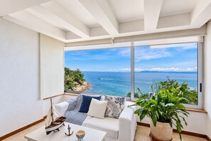 A modern room with a large window overlooking a coastal view, a white sofa with striped cushions, a small table with a ship model and an anchor, and a potted plant.