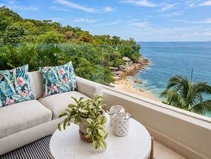 A balcony with a sofa, a potted plant, and a small table with two lanterns, overlooking a beach and ocean.