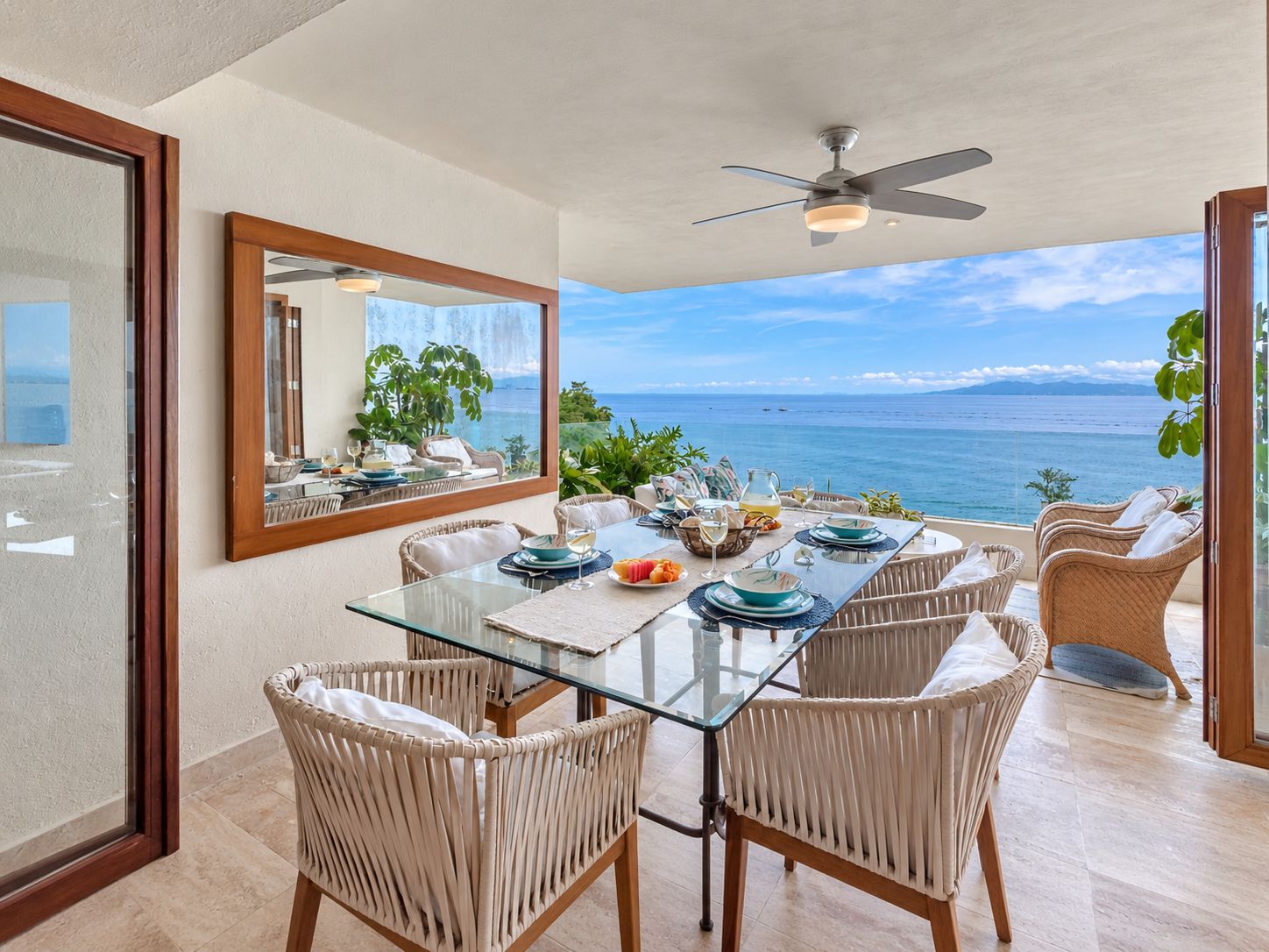 Un coin repas avec une table en verre, des chaises en osier, un ventilateur de plafond et une vue sur la mer.