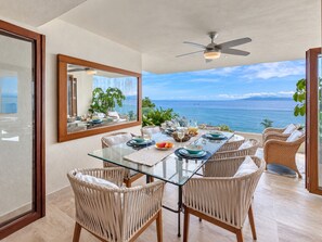 Zona pranzo con tavolo in vetro, sedie in vimini, ventilatore a soffitto e vista sul mare.