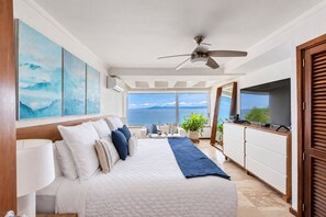 A bedroom with a bed, bedside tables, a ceiling fan, a TV, and a view of the sea.