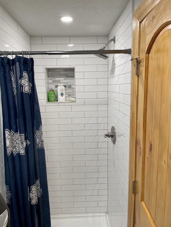 A shower area with white tiled walls, a wooden door, and a shower curtain with a floral pattern. - Cute, cozy, updated cabin in the pines (Pinetop)