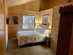 A cozy wooden cabin bedroom with a bed, a window, a lamp, and a painting on the wall.