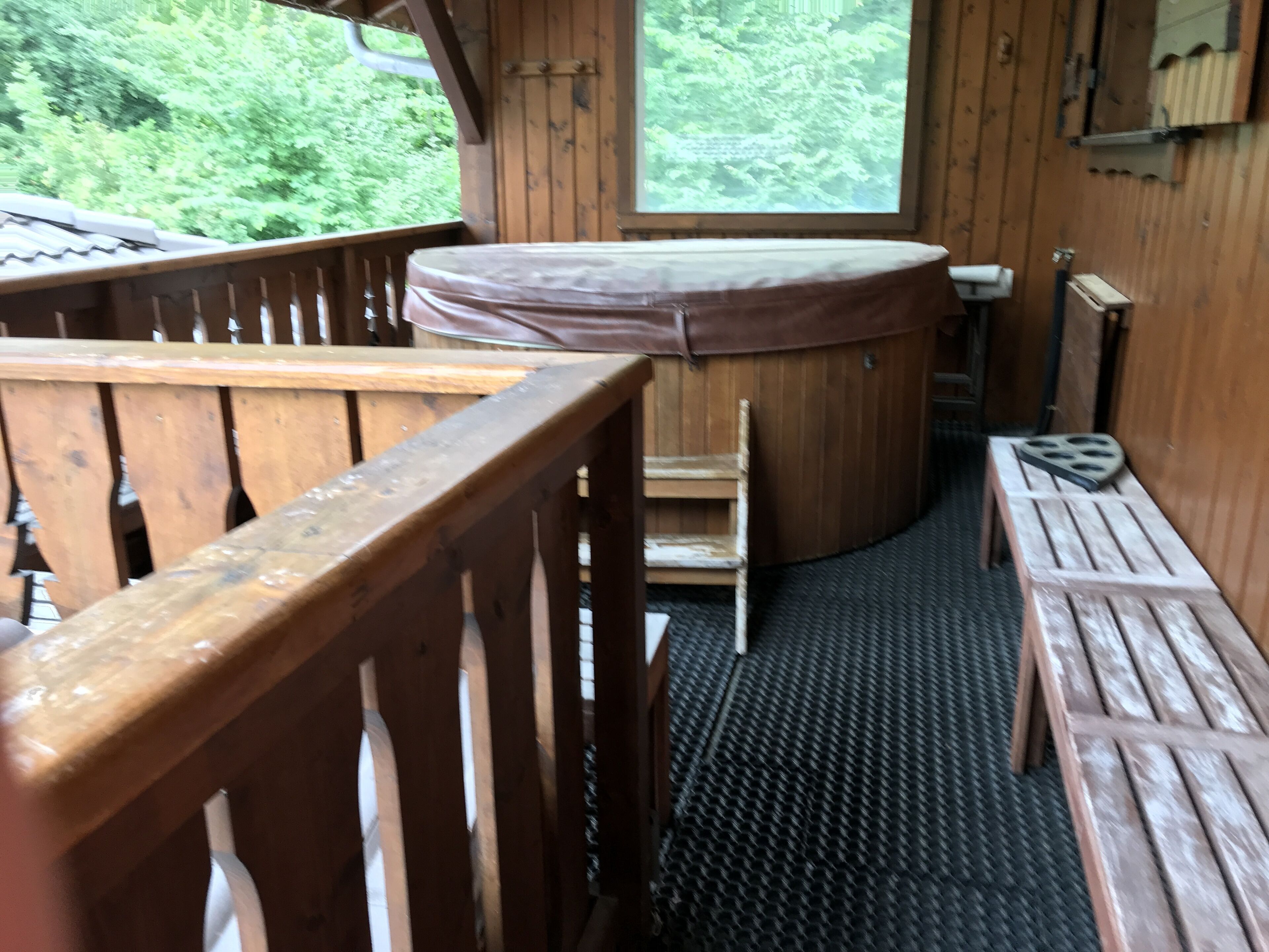 Une terrasse en bois avec un banc et une petite table, une fenêtre donnant sur la verdure et un mur en bois avec une étagère.