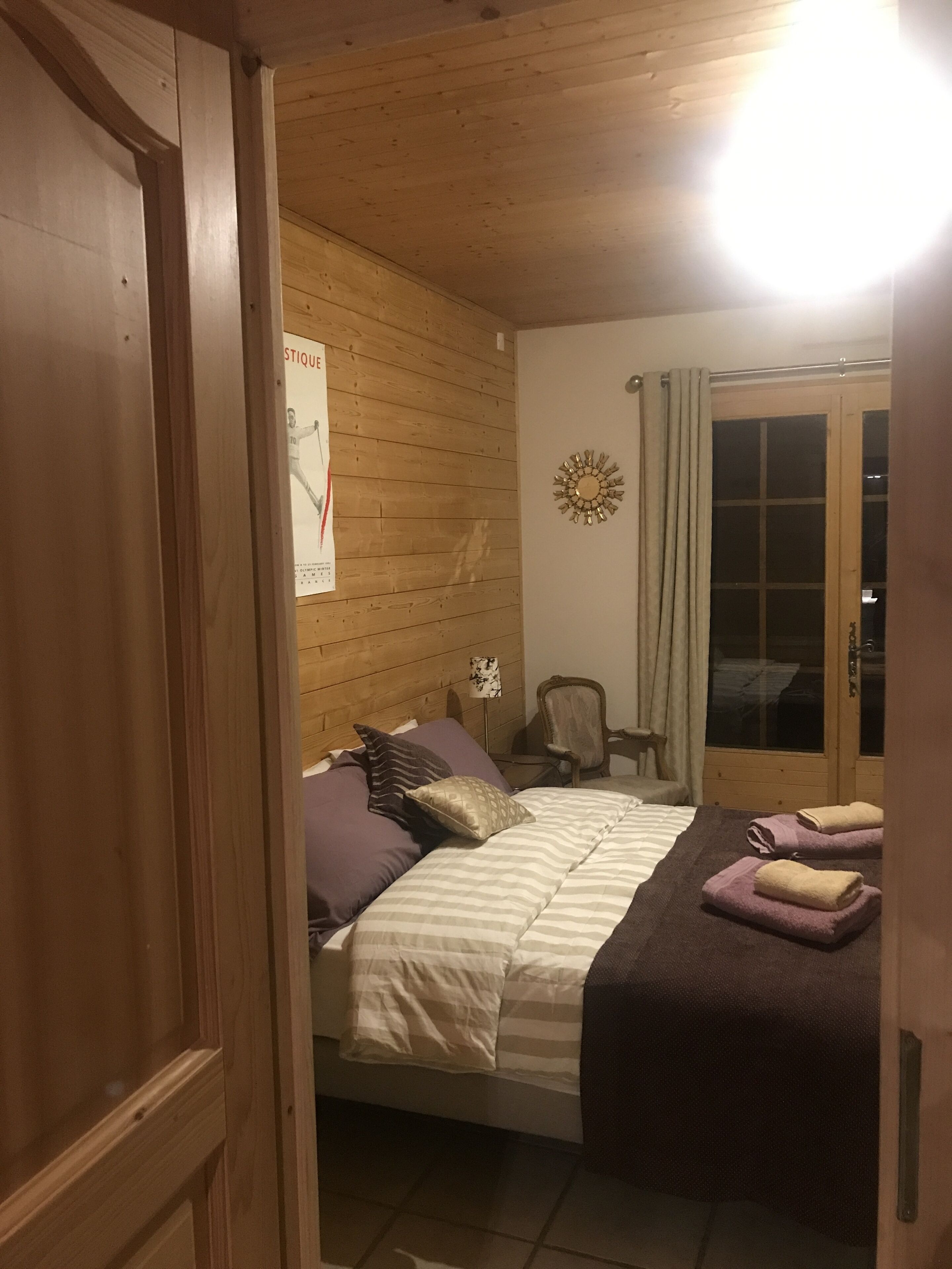 A wooden-paneled room with a bed, a chair, a wall clock, and a window with curtains.