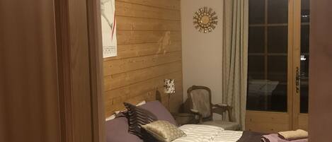 A wooden-paneled room with a bed, a chair, a wall clock, and a window with curtains.