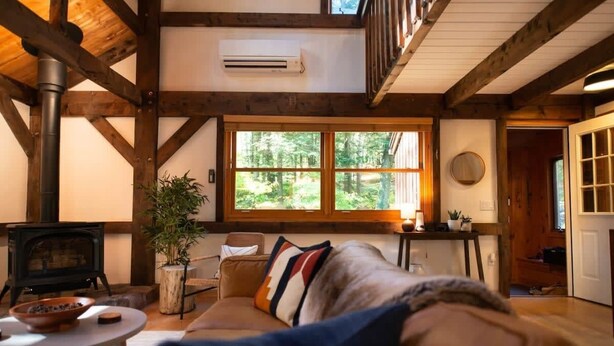 A cozy cabin interior with a wooden ceiling, a fireplace, a sofa, and a large window. - Stonebridge Cabin: Modern Amenities, Enjoy Nature! (Lee)