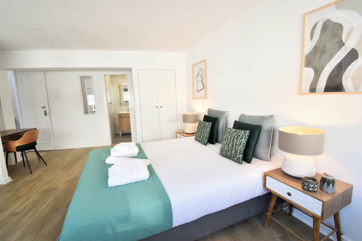 Quarto de hotel com cama, mesa de cabeceira, luminária e escrivaninha com cadeira.