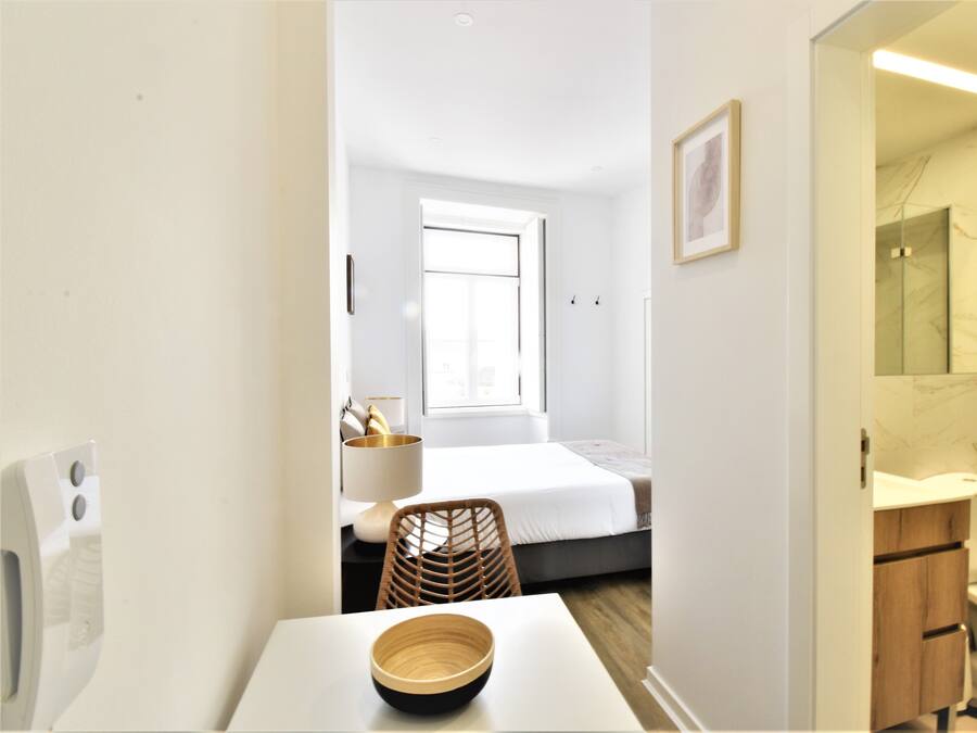 A modern hotel room with a bed, a small table with a bowl, a chair, a window, and a bathroom with a mirror and sink.