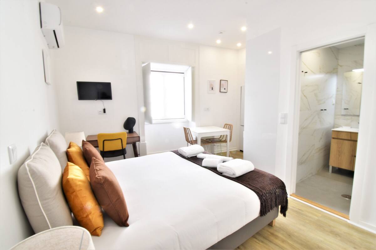 Quarto de hotel com cama, escrivaninha, cadeira, televisão e banheiro visível através de uma porta aberta.