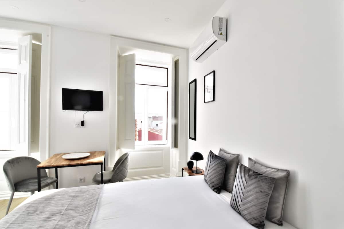 Quarto de hotel moderno com uma cama grande, uma escrivaninha com cadeira, uma televisão de tela plana na parede e uma janela com vista para a cidade.