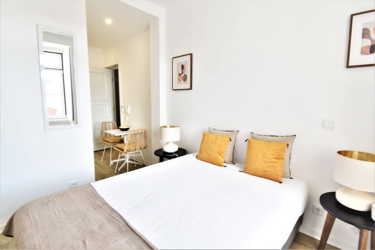 Quarto de hotel moderno com uma cama grande, uma área de estar com uma cadeira e mesa, e um quadro na parede.